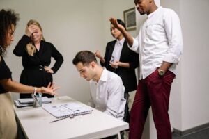 Foto de um homem sentado na mesa do escritório, de cabeça baixa, submisso, enquanto outras pessoas em pé a sua volta parecem estar lhe dando bronca e humilhando. A imagem ilustra o artigo