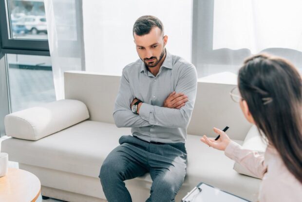 Homem sentado em sofa branco de braços cruzados e ao lado uma psicanalista conversando com ele. A imagem ilustra o destaque 5 da Home do Site Online Psicanalista.